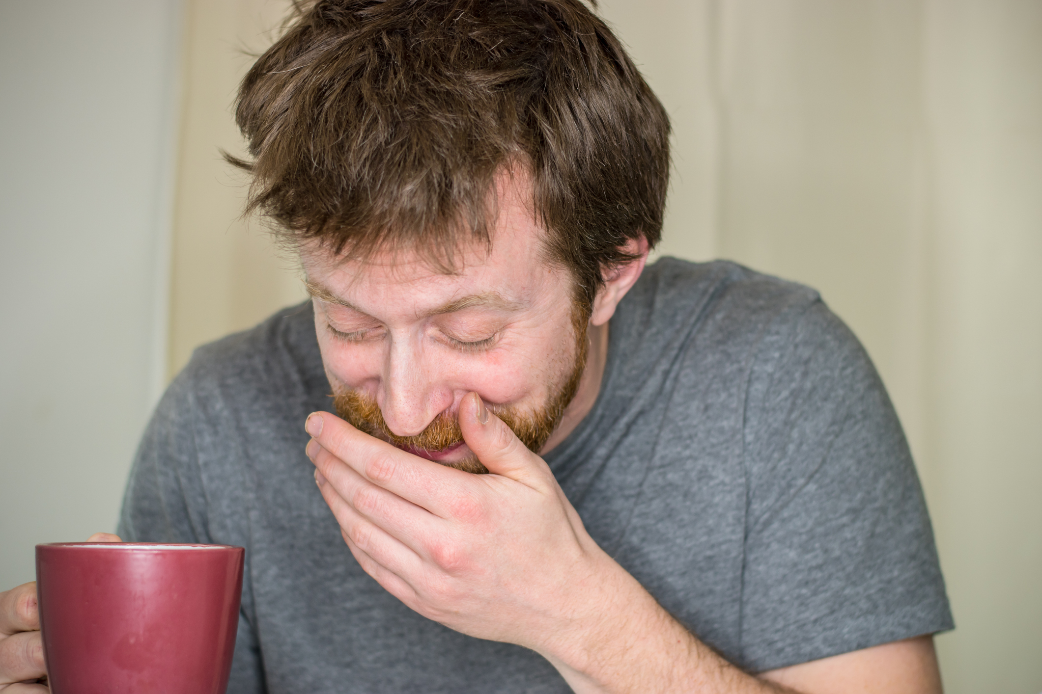 Man laughs while holding a mug, hand covering his mouth