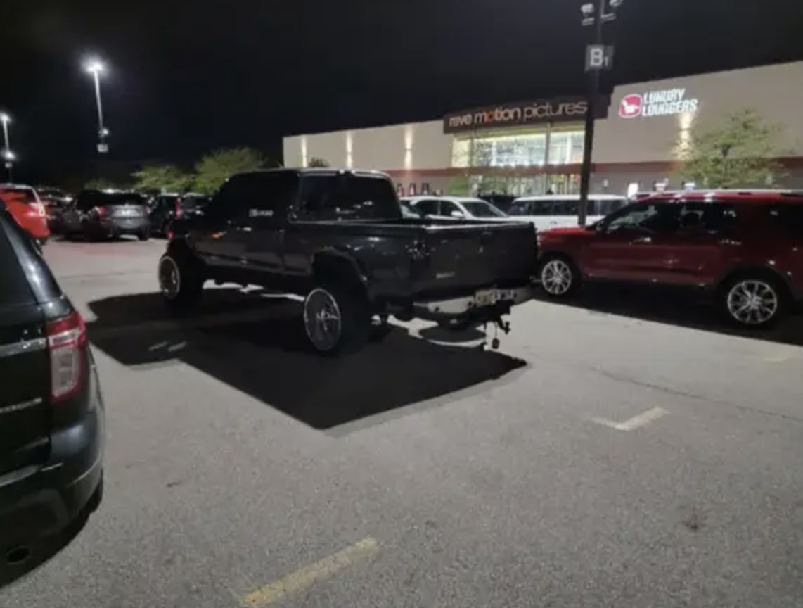Pickup truck with oversized wheels parked across multiple spots at night outside a movie theater