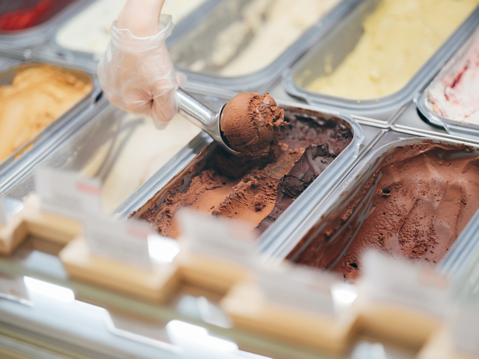 Person scooping chocolate ice cream from a display case, suggesting a work scene at an ice cream shop
