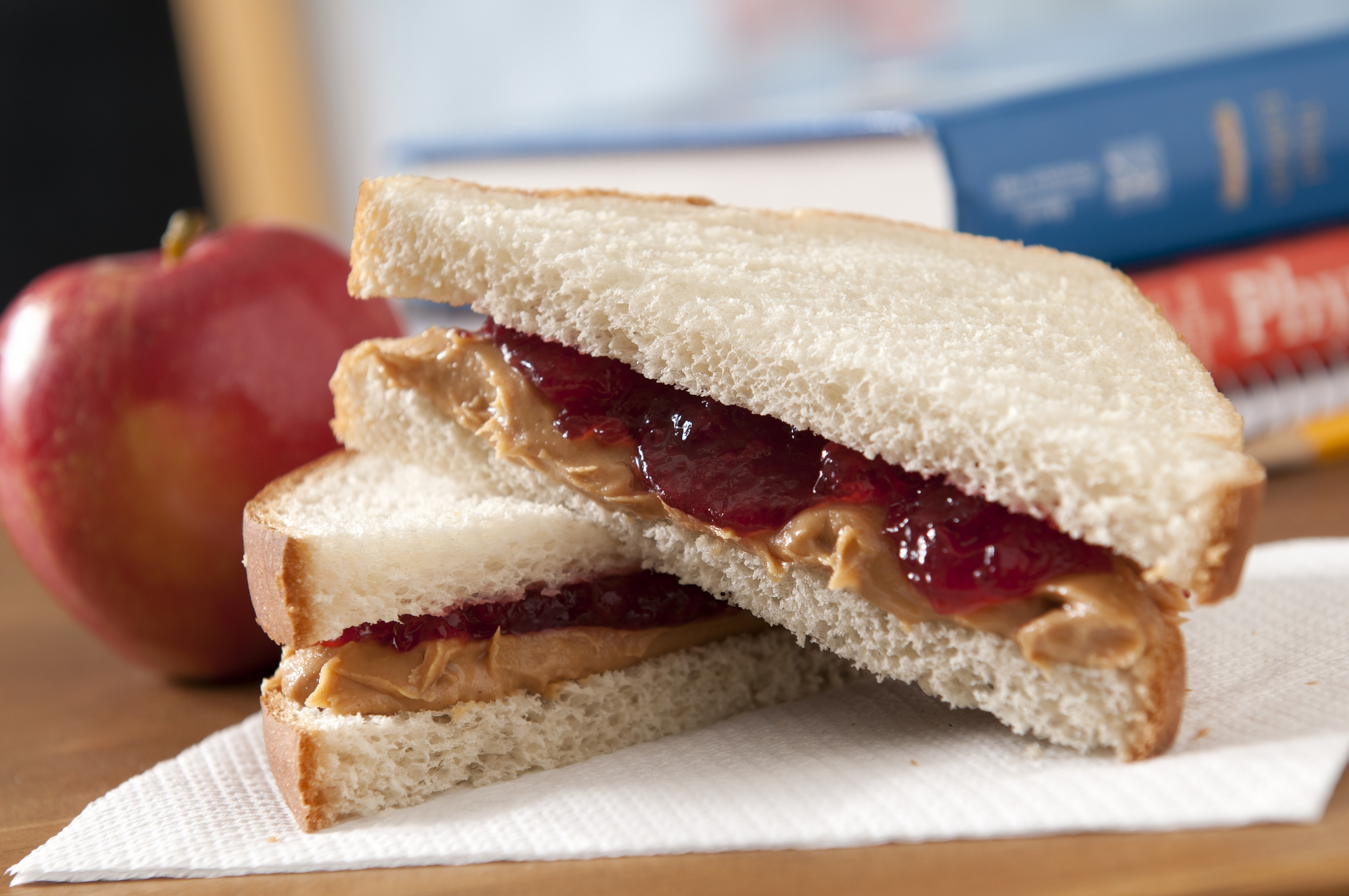 Peanut butter and jelly sandwich on white bread, with a whole apple and books in the background