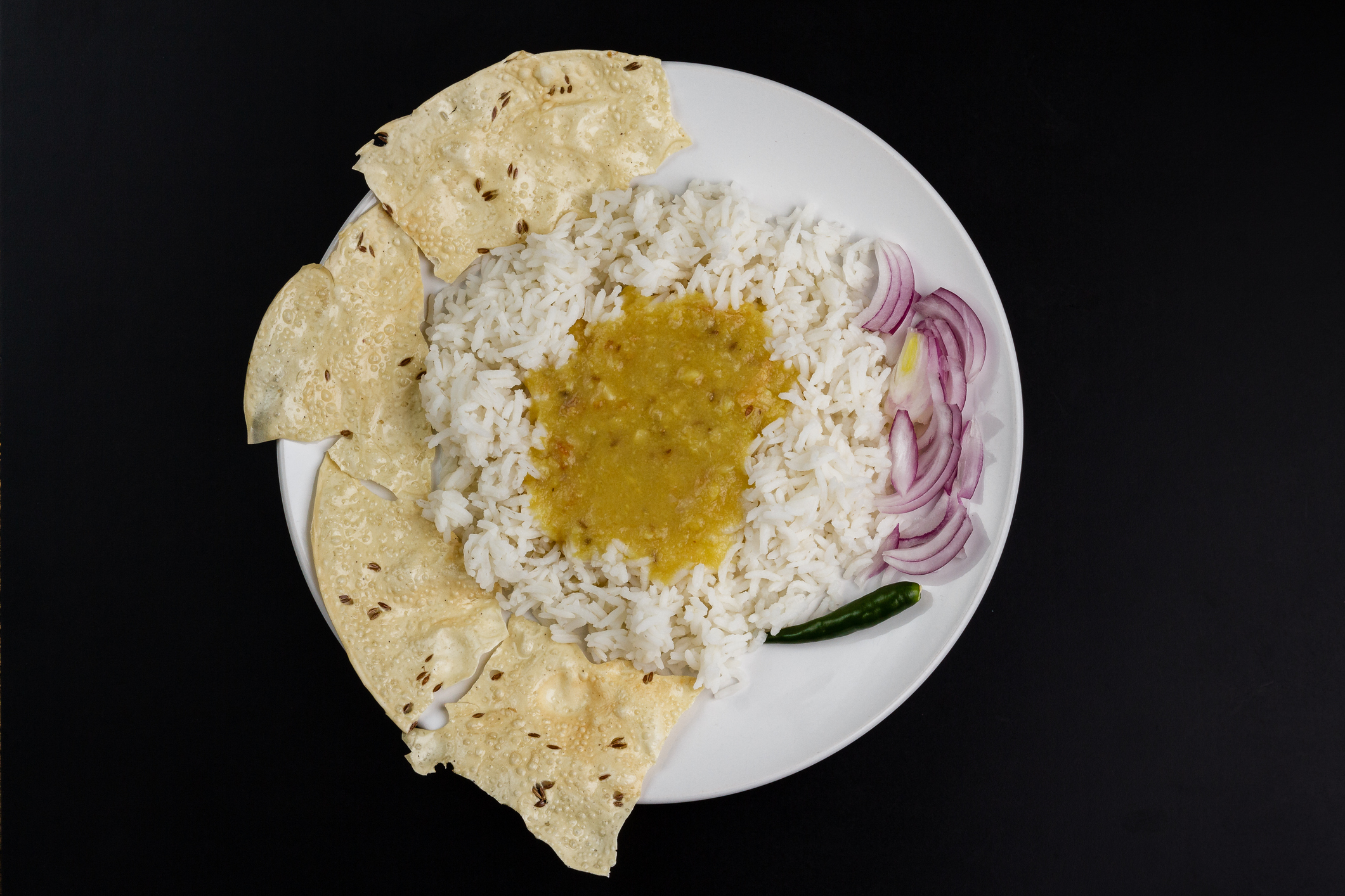 A plate with rice, lentil curry, papadum, a green chili, and sliced onions