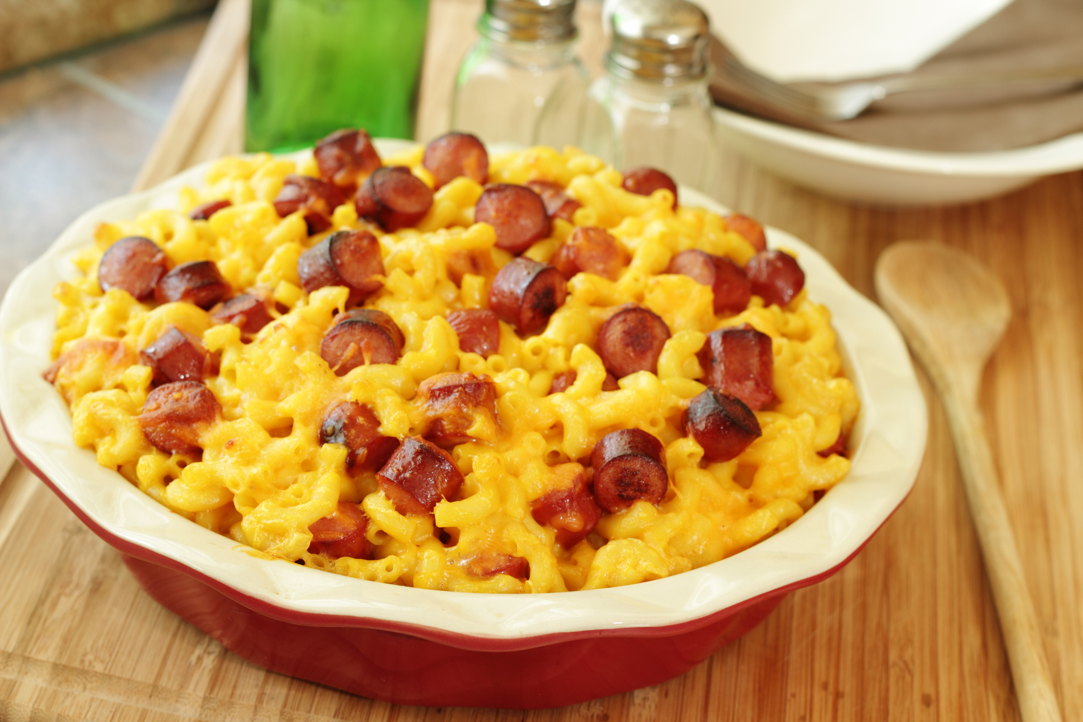 A dish of macaroni and cheese with sliced hot dogs mixed in, served on a wooden table