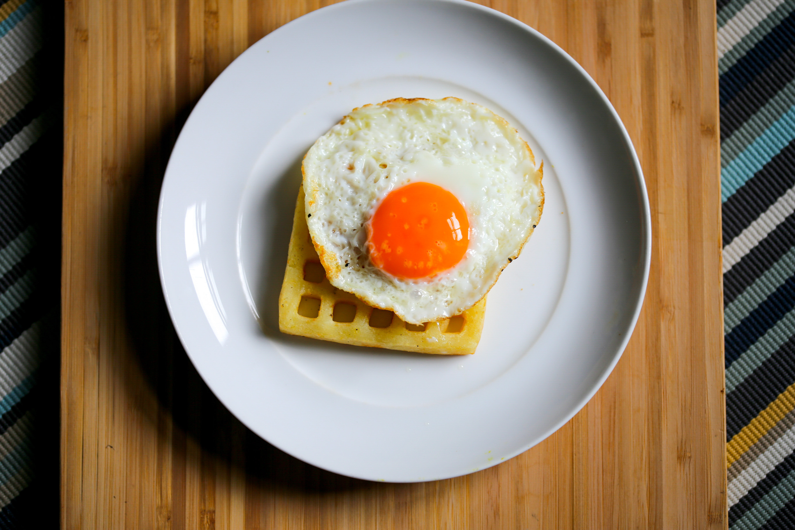 A fried egg atop a waffle on a white plate, served on a striped placemat