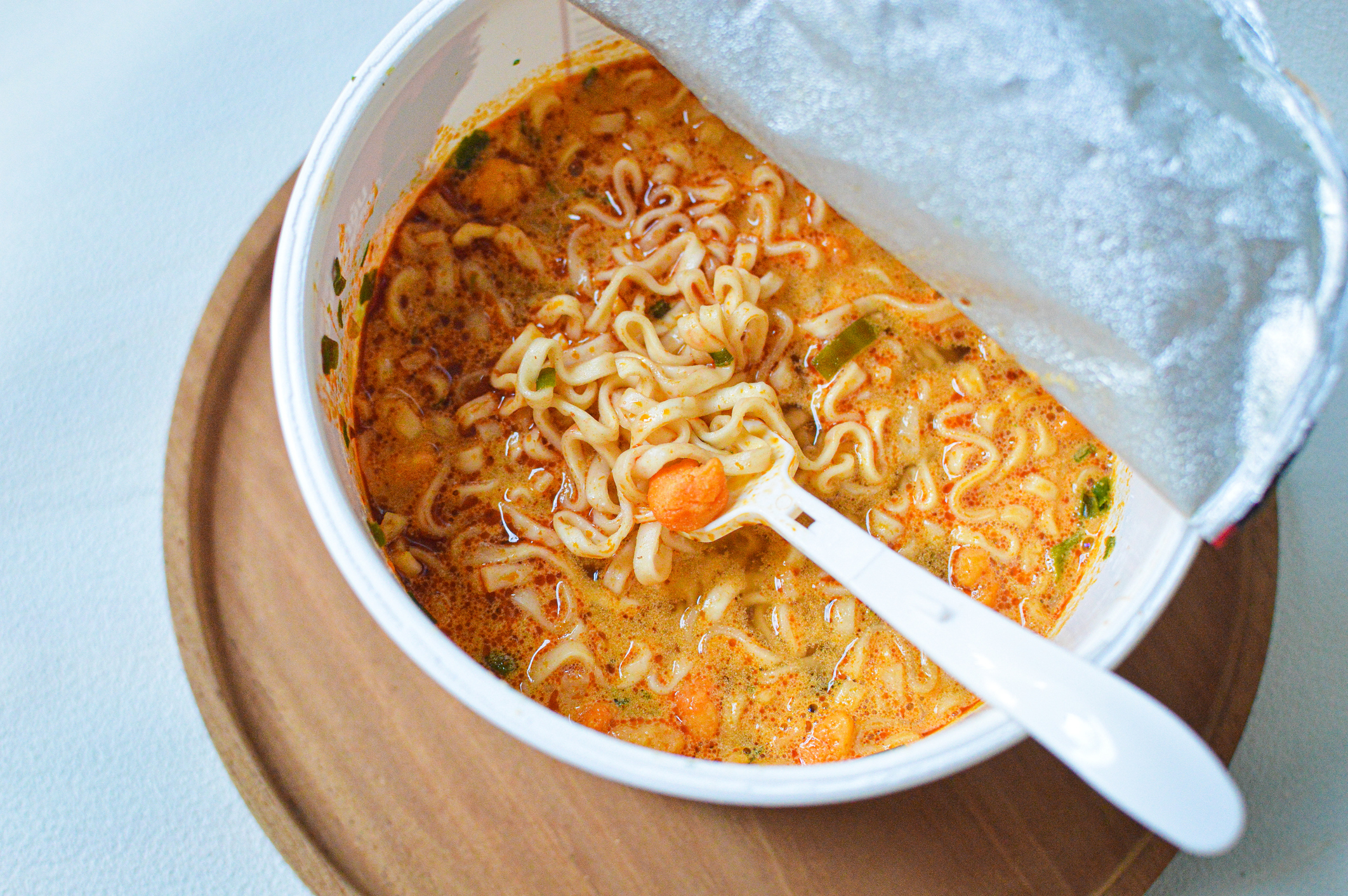 Bowl of spicy instant noodles with vegetables, partially covered by lid, with a spoon