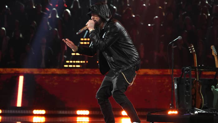 Musician in leather jacket and hat performing onstage with microphone, background has red lighting and audience silhouette