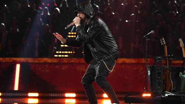 Musician in leather jacket and hat performing onstage with microphone, background has red lighting and audience silhouette