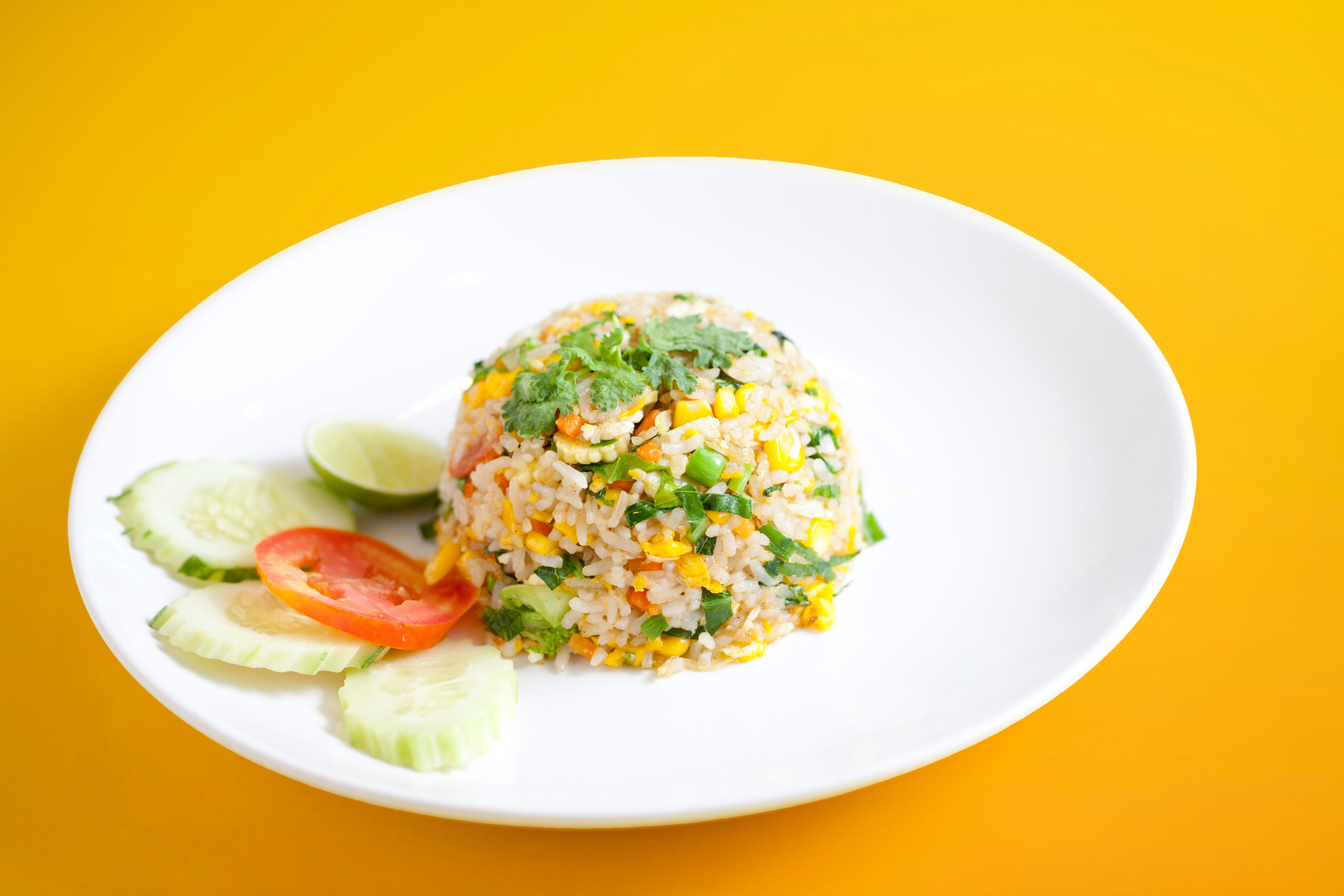 A plate of fried rice with herbs, lime wedges, and slices of cucumber and tomato on a yellow background