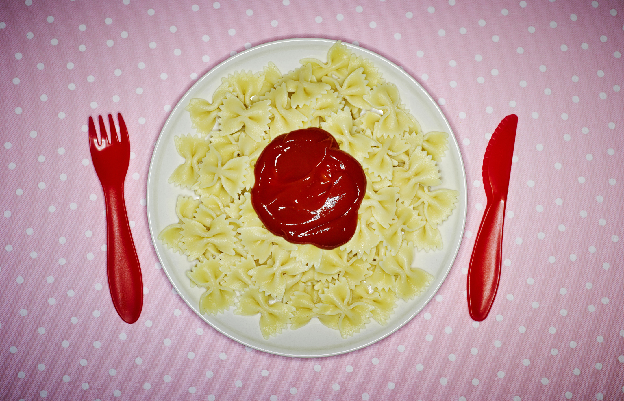 Plate of pasta with a dollop of sauce, flanked by plastic fork and knife on a dotted surface