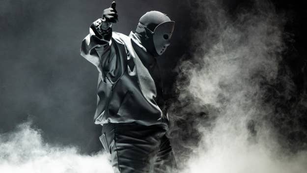 Music artist in mask and hoodie on stage with dramatic lighting and smoke, pointing upwards