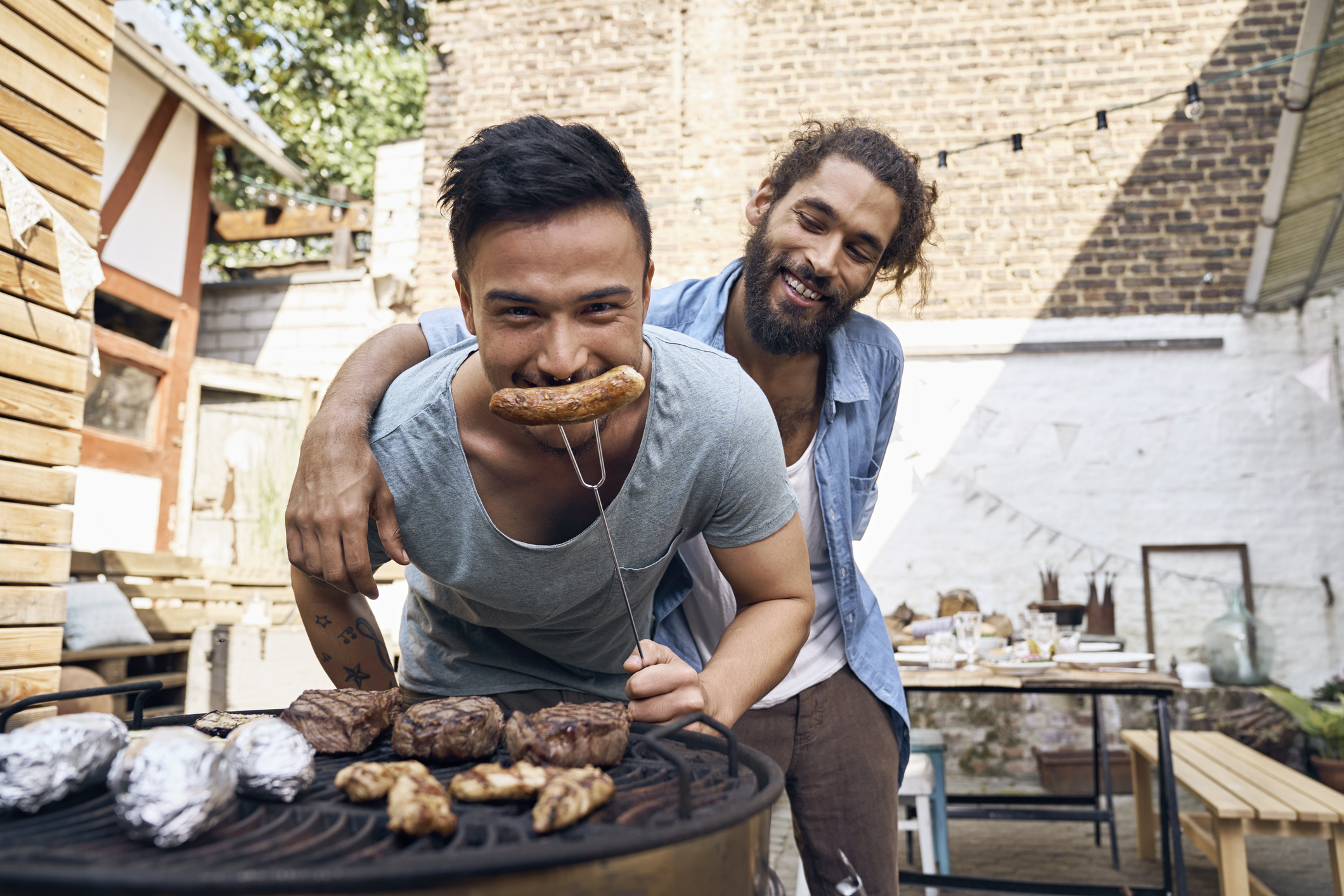Two men enjoying a barbecue, one biting into a grilled sausage, smiling and bonding in a casual outdoor setting
