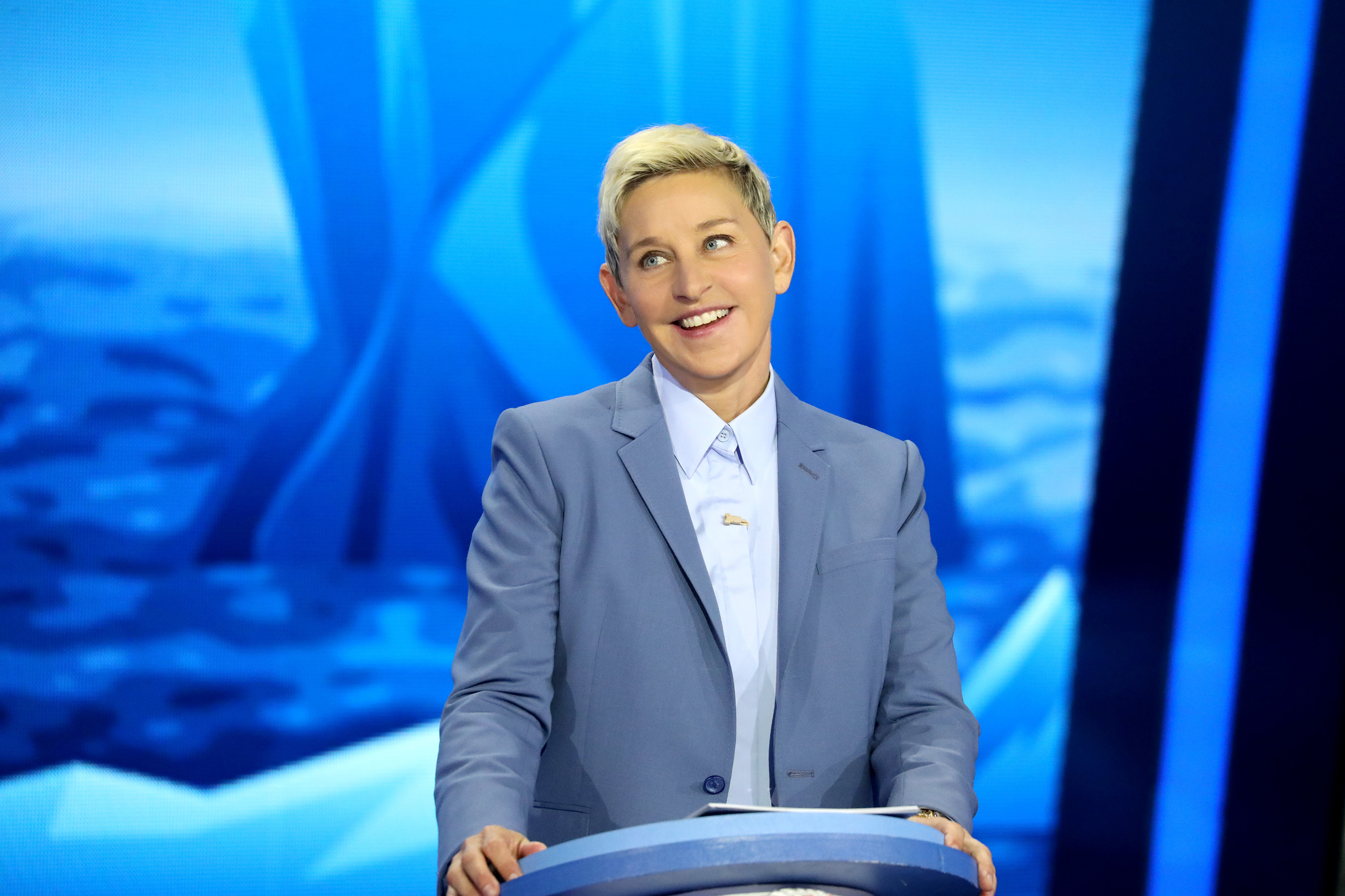 Ellen DeGeneres on a TV set, smiling, in a blue suit and white shirt, standing behind a podium