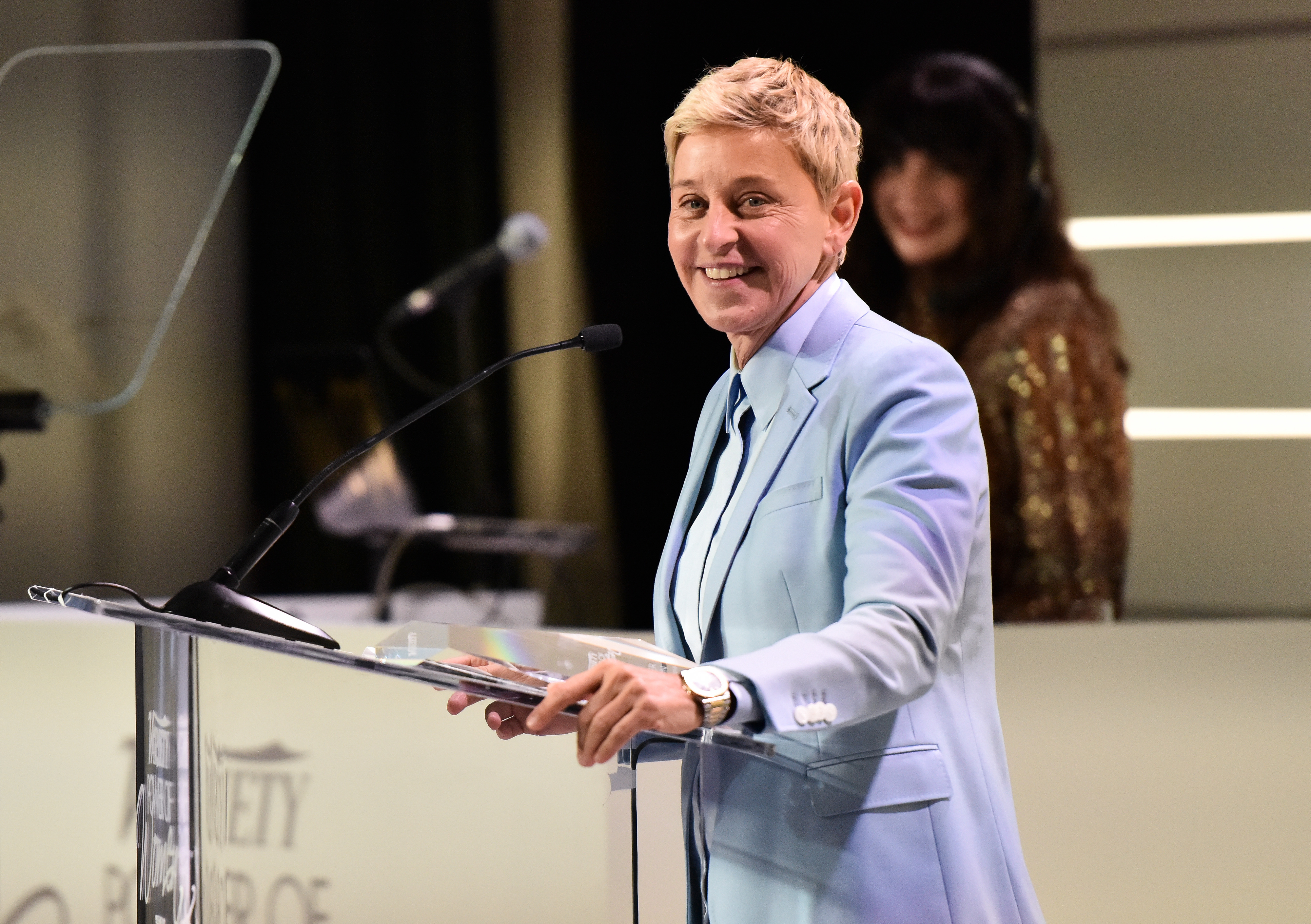 Ellen DeGeneres in a light blue suit speaking at a podium with a smile, another person in background