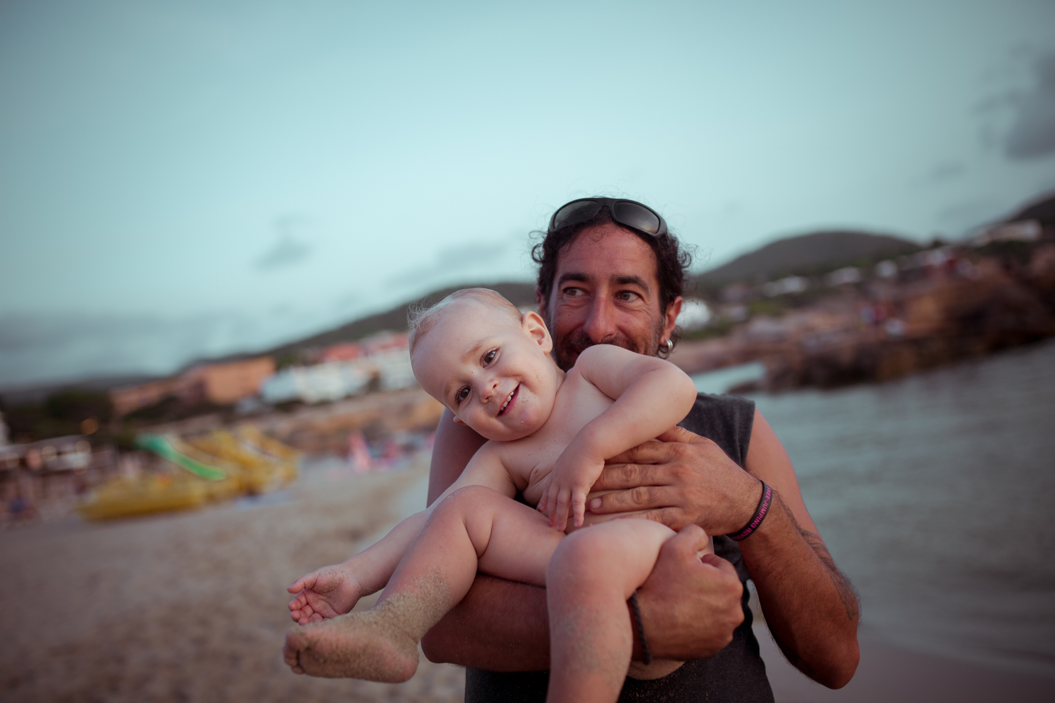 Man holding a smiling baby at a beach, both looking at camera, with coastal landscape in the background