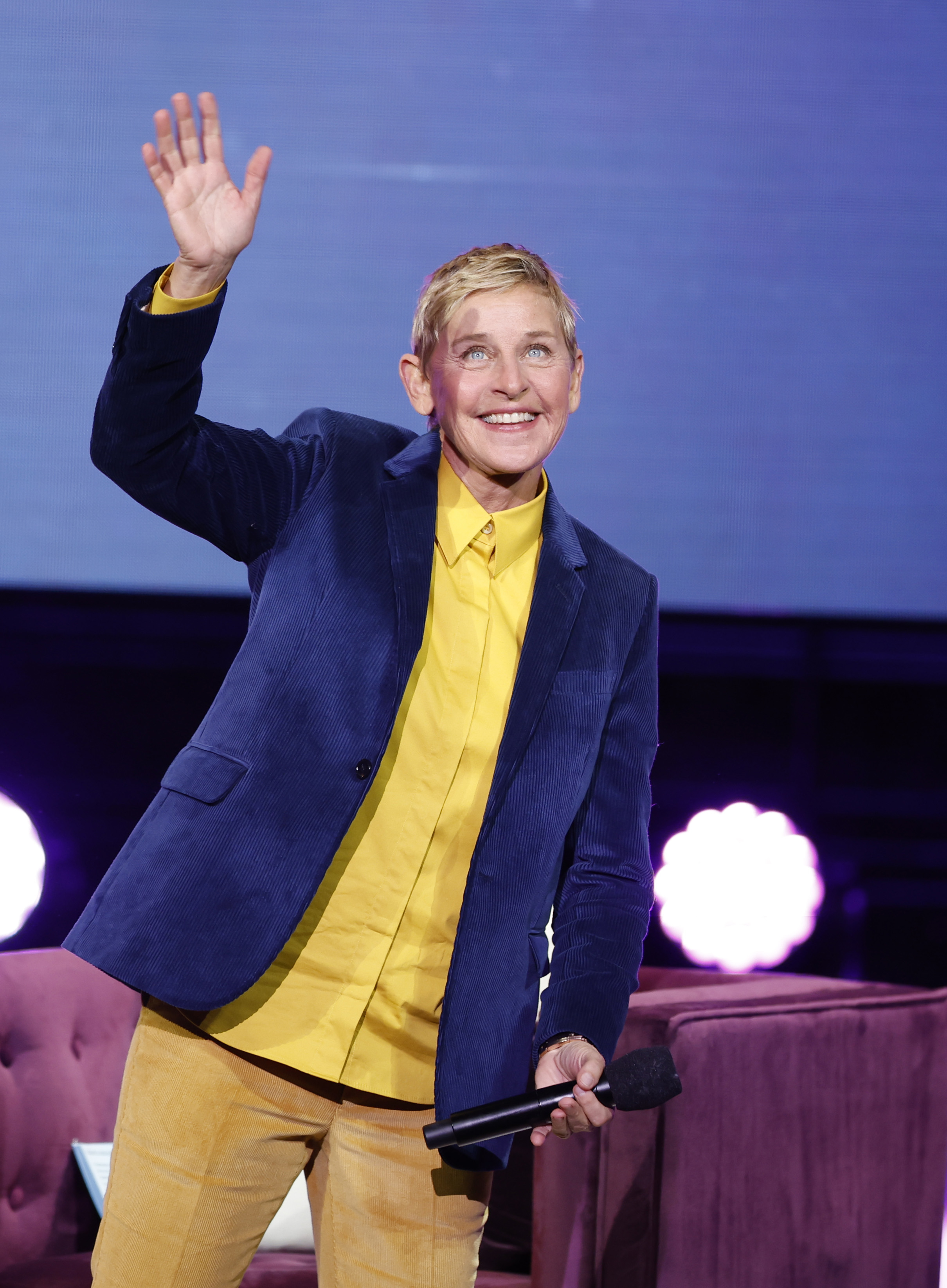Ellen DeGeneres on stage in a blazer and trousers waving, with a mic in hand