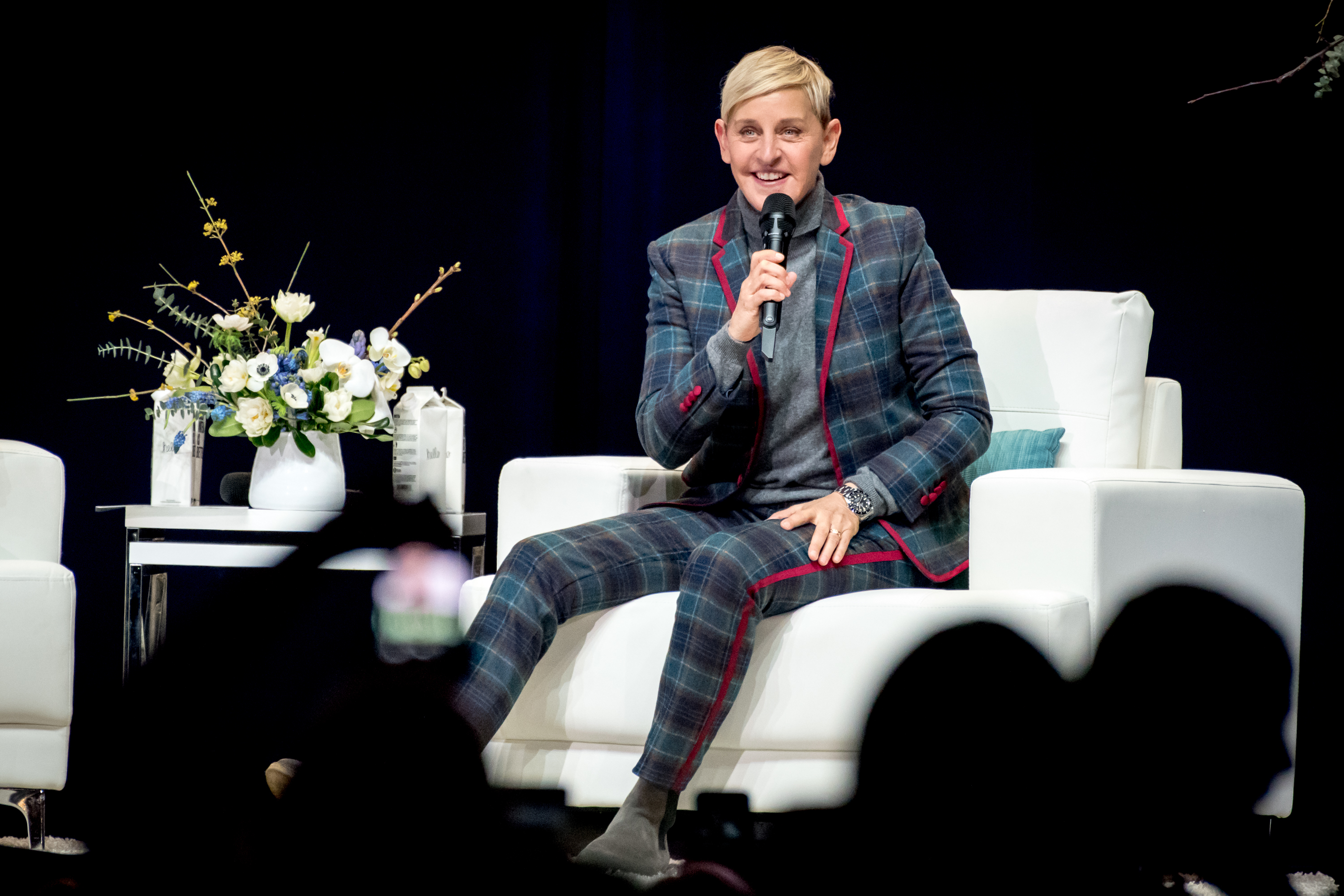Ellen DeGeneres speaking at an event, wearing a plaid suit and seated on a white chair onstage
