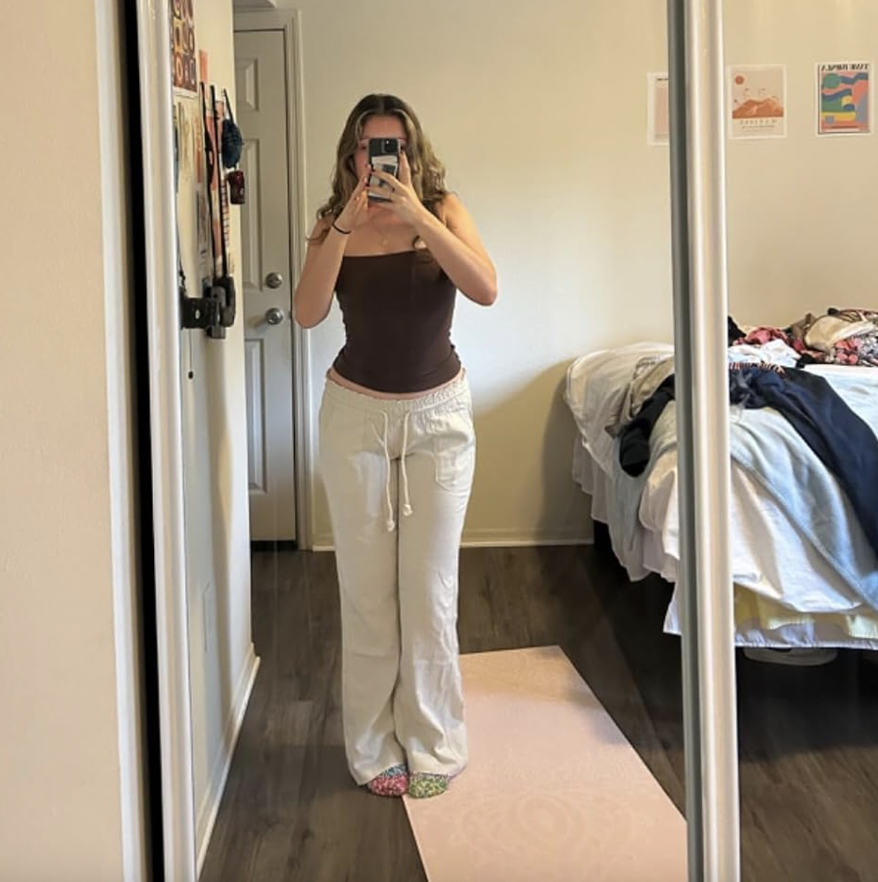 Person in mirror taking photo with phone, wearing a tank top and white pants, messy bed in background