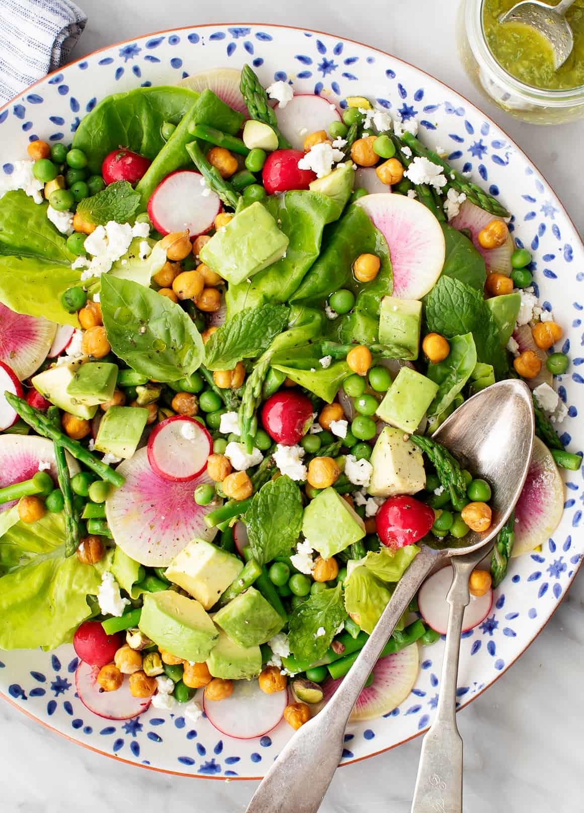 A fresh salad with lettuce, chickpeas, avocado, radishes, peas, and cheese, served with dressing and utensils on the side