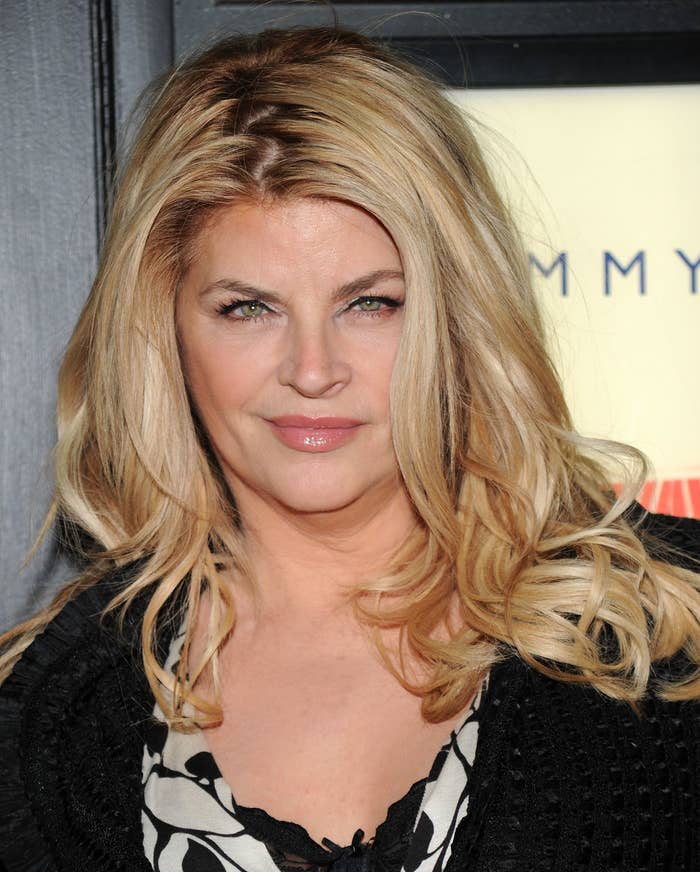 Kirstie Alley wearing a black knit cardigan and floral dress, posing at an event