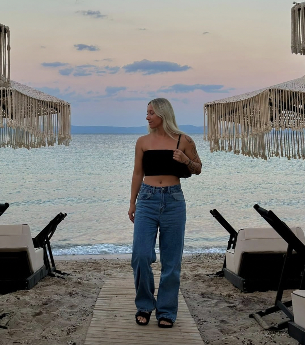 Woman in a casual black top and blue jeans standing on a beach boardwalk at dusk