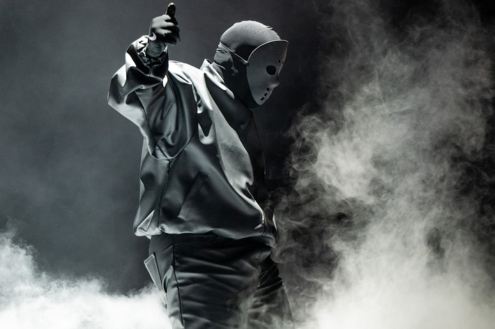 Music artist in mask and hoodie on stage with dramatic lighting and smoke, pointing upwards