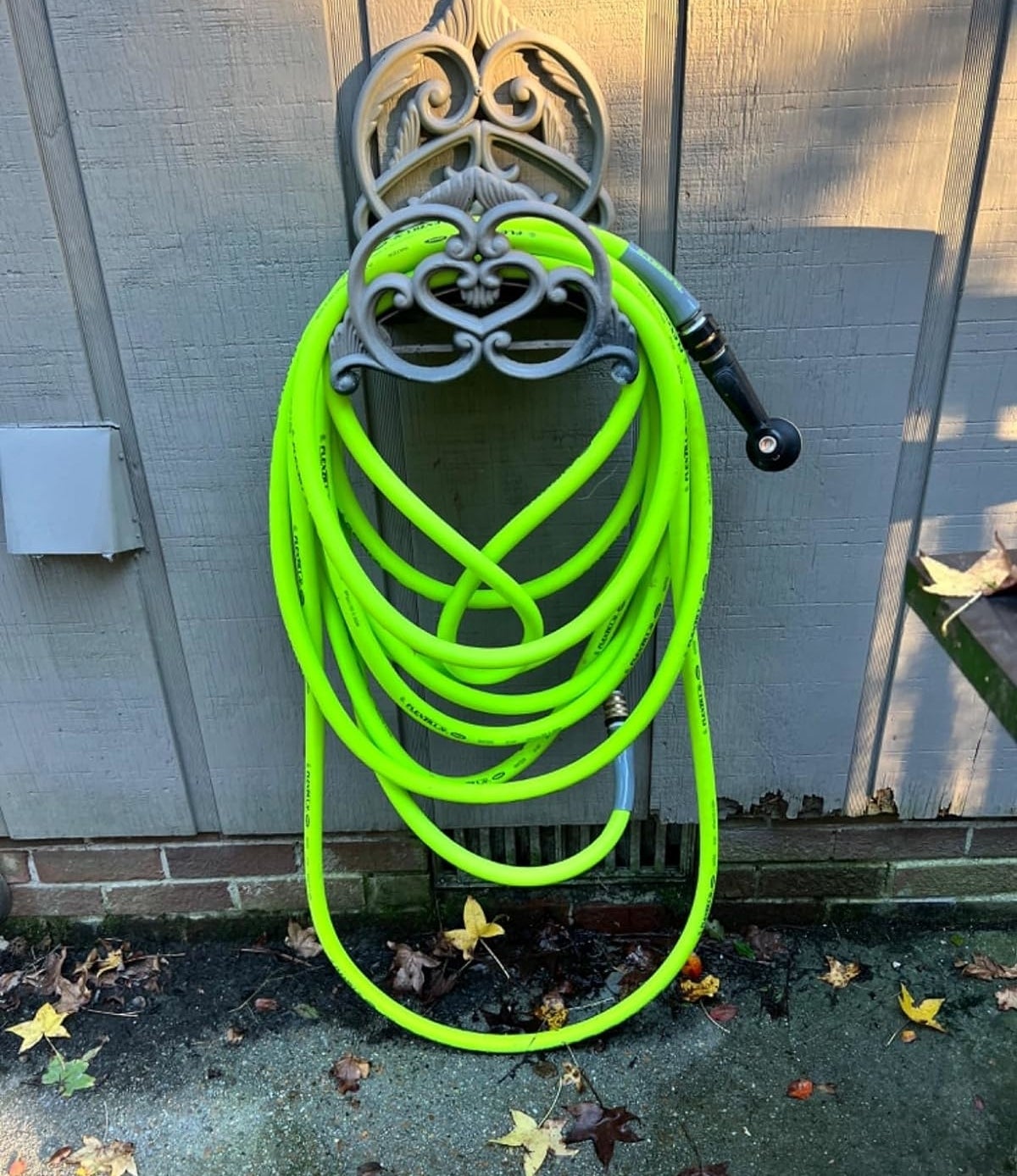 A green garden hose coiled around a decorative wall-mounted holder with fallen leaves on the ground