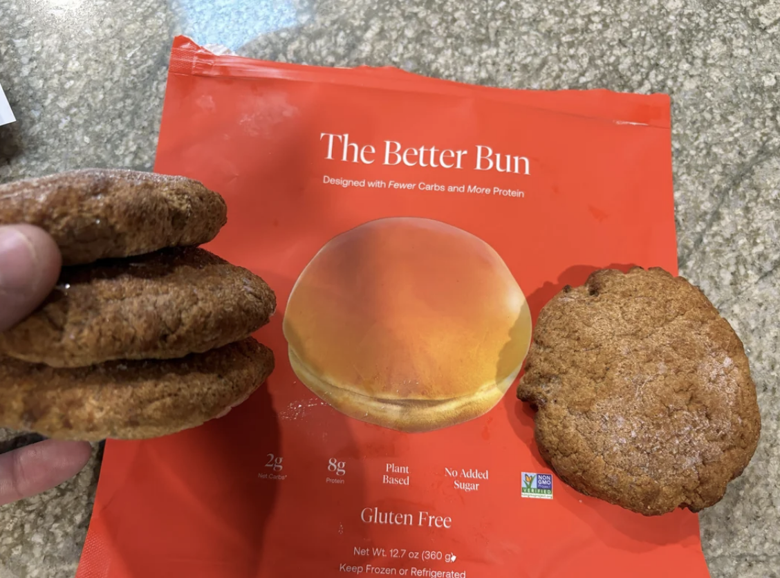 A hand holds two burger buns above a package labeled &#x27;The Better Bun&#x27;, highlighting the product&#x27;s nutritional benefits
