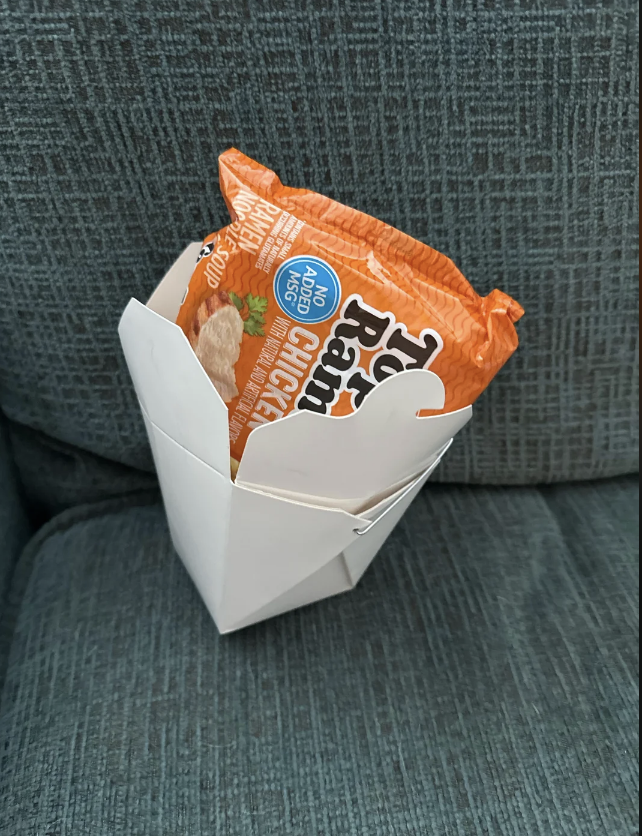 A bag of potato chips propped open inside a white takeout container on a couch