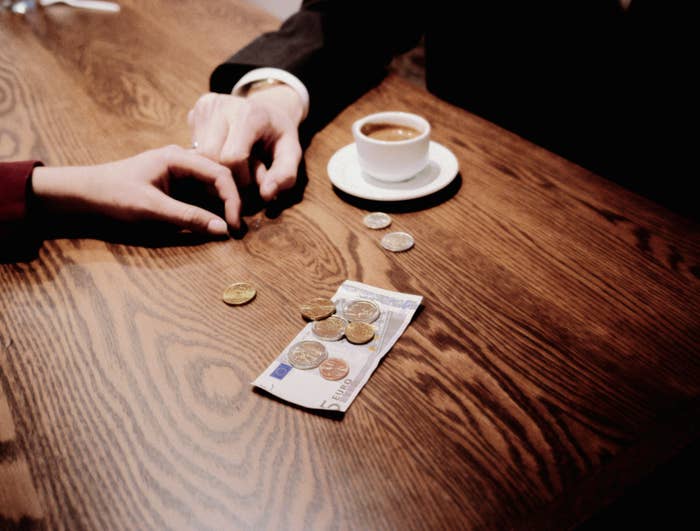 Two people&#x27;s hands on a table, one placing a coin in another&#x27;s hand, with a coffee cup and money visible