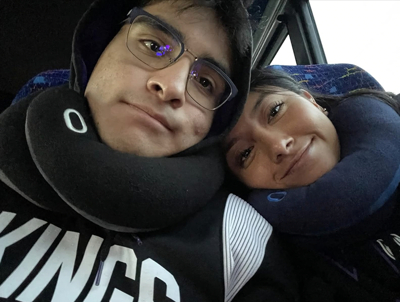 Two people with travel pillows on a bus, one wearing glasses and a hoodie, the other leaning on their shoulder