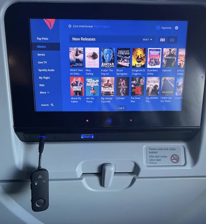 In-flight entertainment screen showing various movie options, headphones plugged in, and &#x27;Fasten seat belt&#x27; sign illuminated above