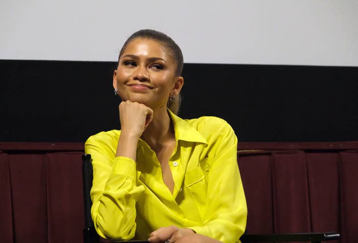 Zendaya in a bright blouse, resting chin on hand, smiling during an event