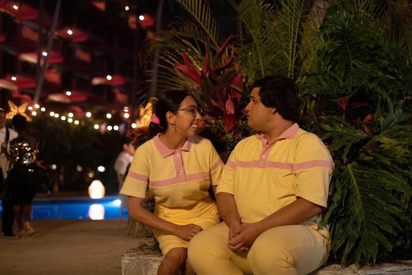 Lorena y Memo, personajes de la serie Acapulco, sentados juntos en una banca, luciendo polos a juego y pantalones color crema, mirándose el uno al otro