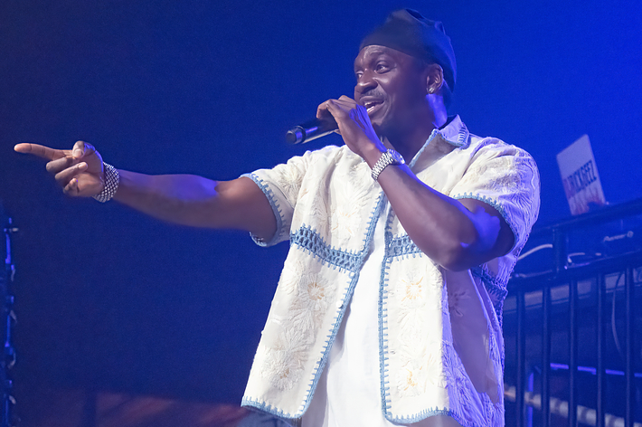 Pusha T performing on stage, pointing outwards, wearing a traditional styled top, ambient stage lighting visible