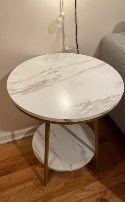 Round marble-patterned side table with a lower shelf, near a grey couch and against a beige wall