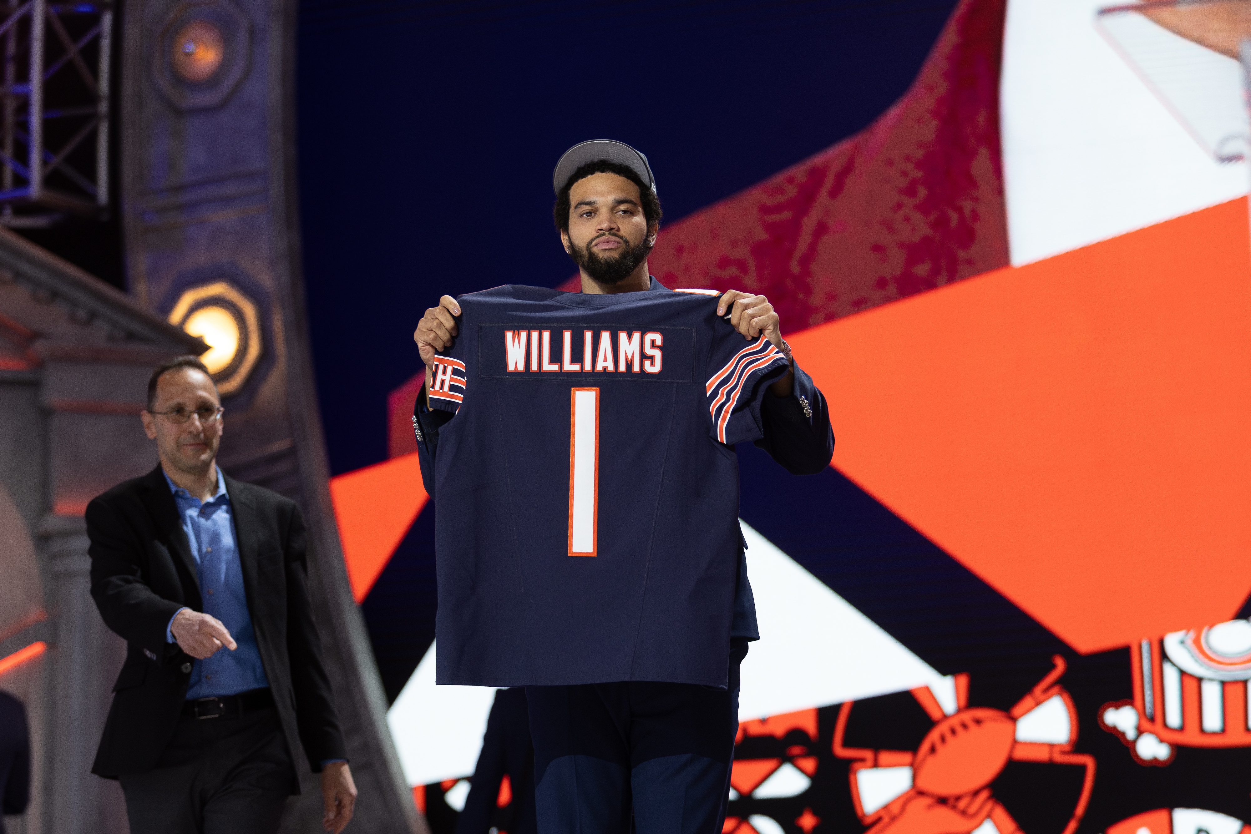 Person on stage holding a sports jersey with &quot;WILLIAMS 1&quot; on it at a draft event, with a graphic backdrop and another individual nearby
