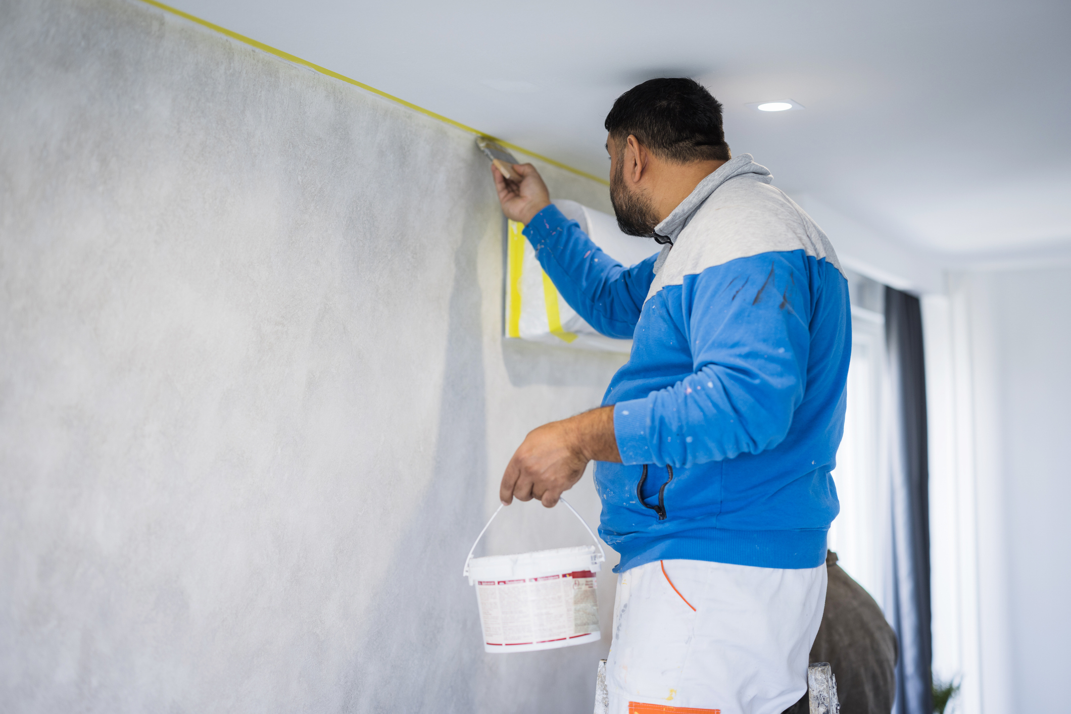 Person applying tape to a wall in preparation for painting, holding a paint bucket