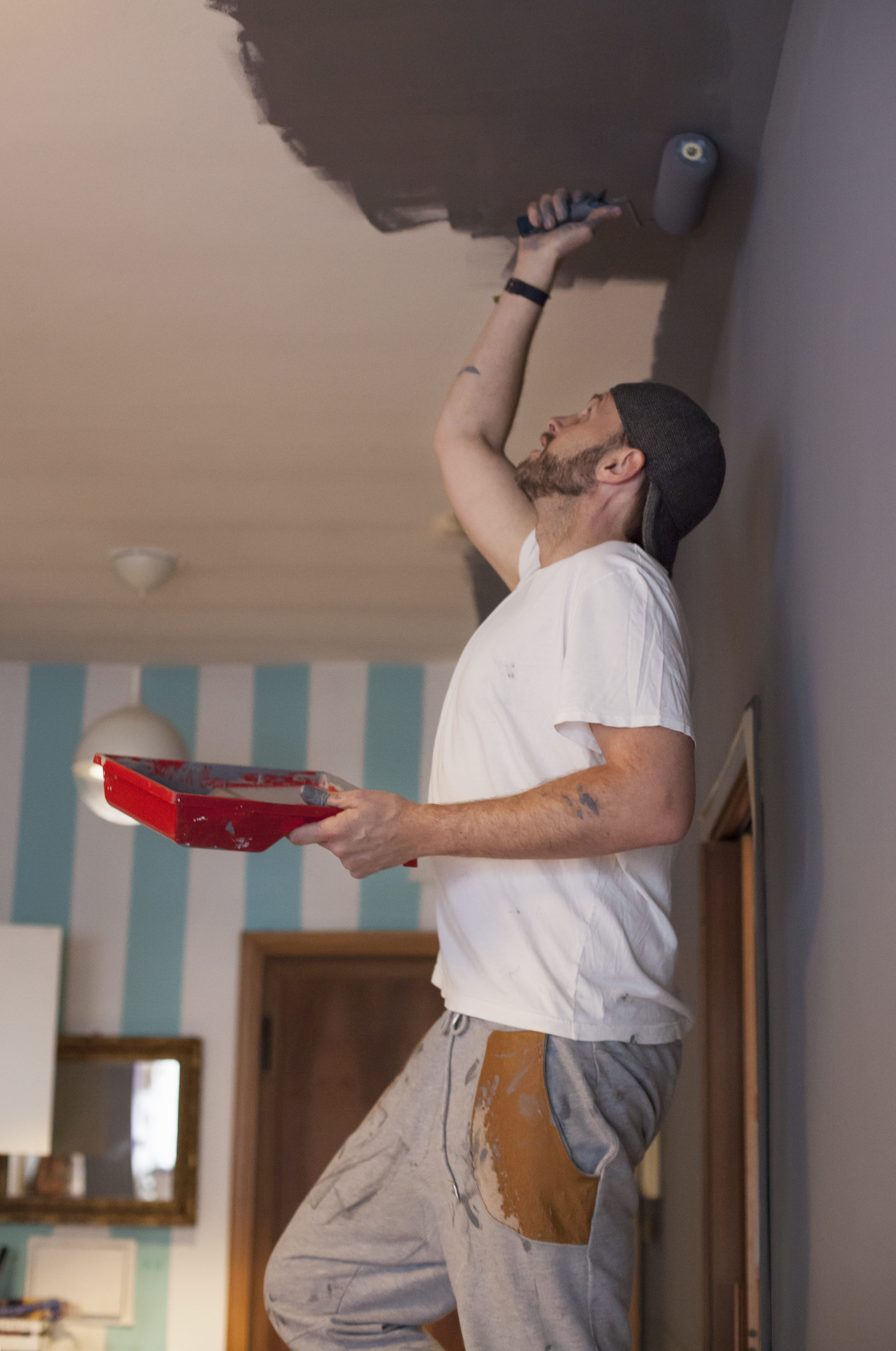Man painting ceiling with roller, wearing a white shirt and speckled pants, indoors