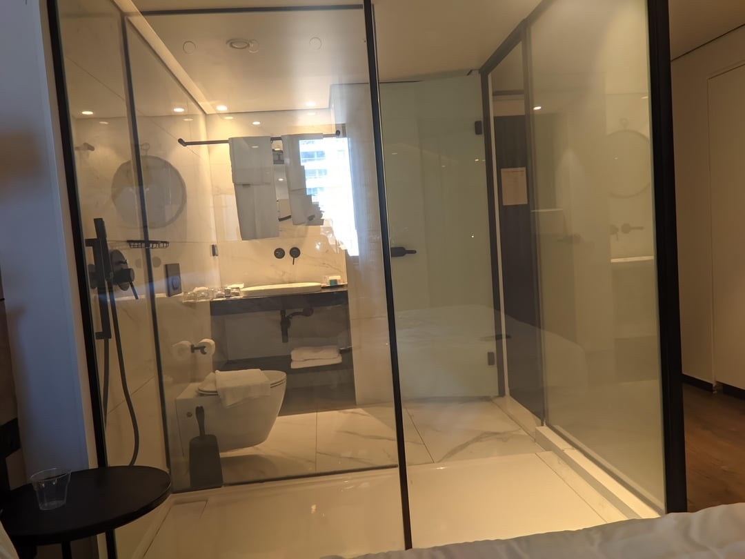 View of a hotel room&#x27;s interior looking into a glass-walled bathroom with a visible toilet and shower