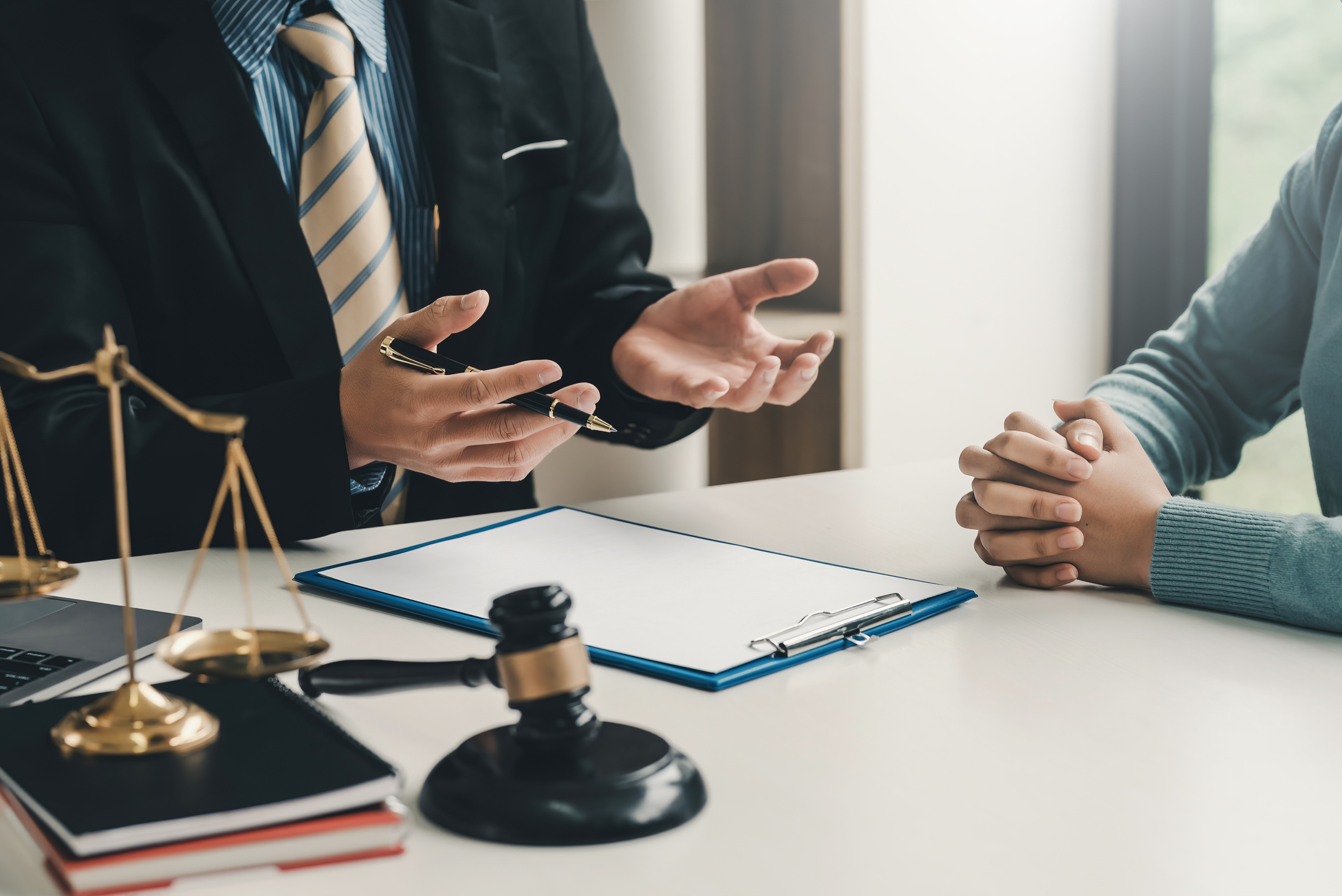 A lawyer in a suit gestures while talking to a client across a desk with legal documents and a gavel present