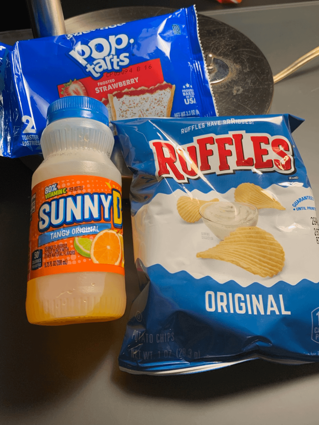 A bottle of SunnyD, a pack of strawberry Pop-Tarts, and a bag of original Ruffles chips on a desk