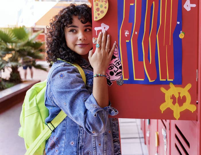 Joven sonriente apoyada en un casillero con pegatinas, lleva chaqueta de mezclilla y mochila