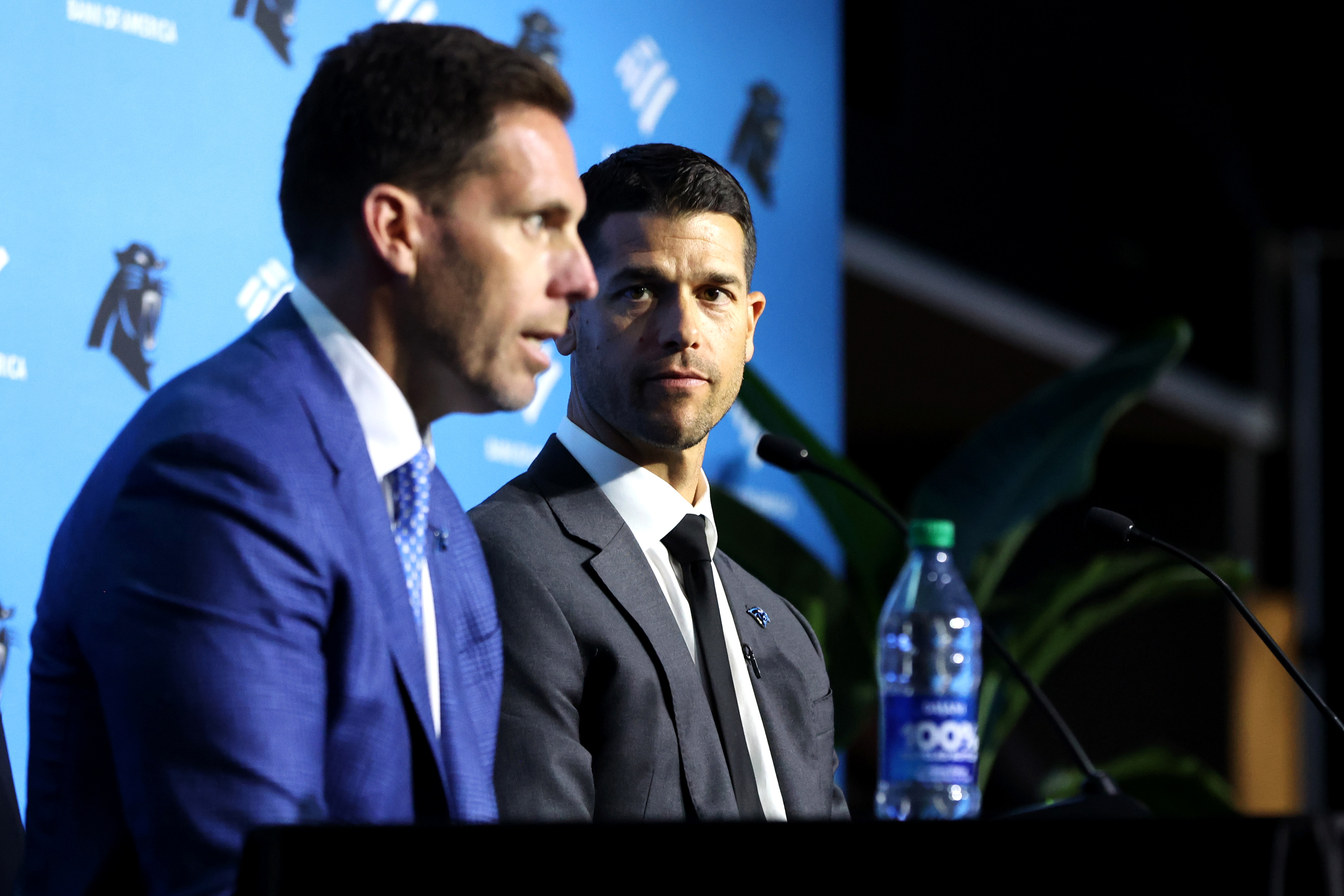 Two men in suits at a sports press conference, one speaking at the podium