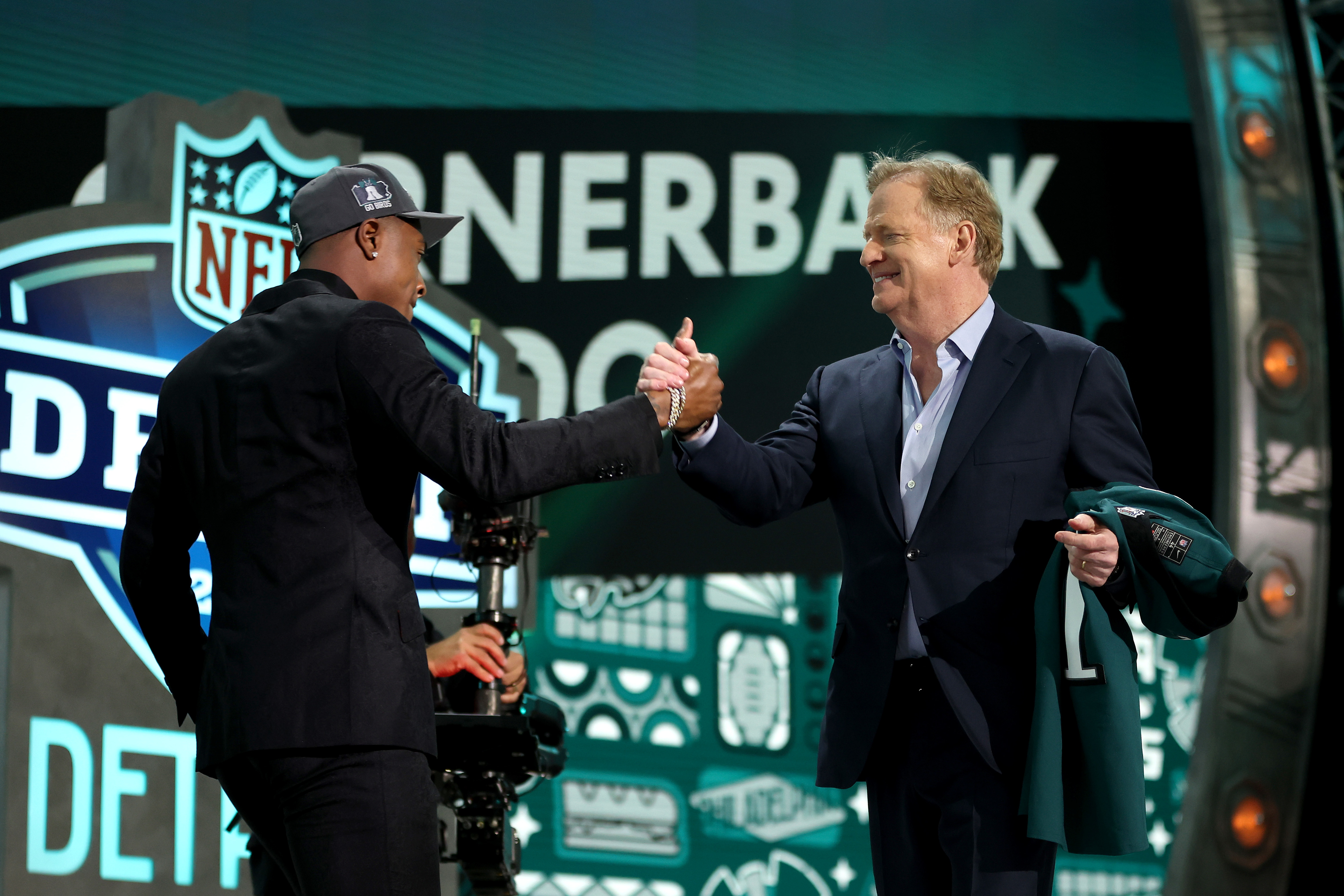 Two men on stage at an NFL event shaking hands, one passing a cap, backdrop with NFL logos