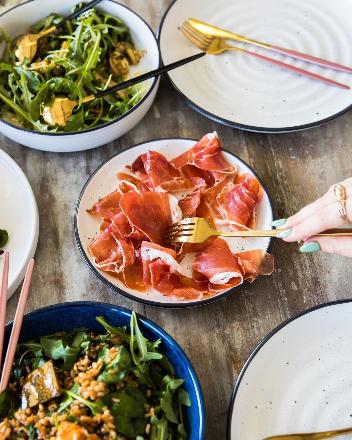 Person dining on prosciutto and salad at a table with multiple dishes