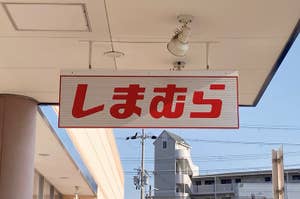 Sign with Japanese text "しまむら" indicating the entrance of a Shimamura store, with buildings in the background