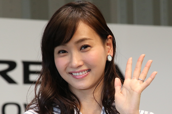 画像にはストライプのシャツを着た女性が笑顔で手を振っています。