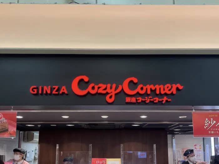 Storefront sign for "GINZA Cozy Corner" with two employees wearing masks standing behind counters
