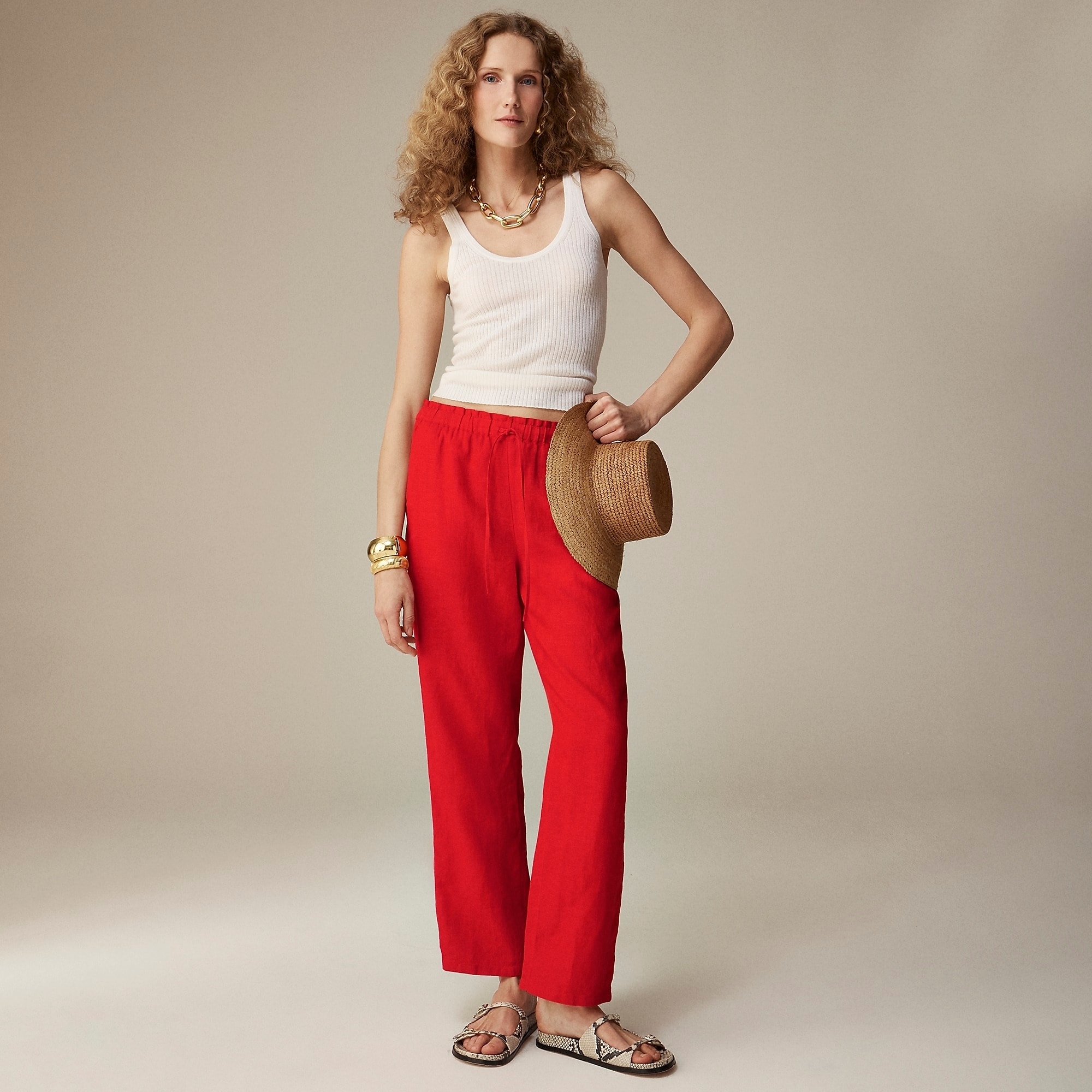 Woman in a white tank top, red trousers, and sandals, holding a straw hat, posing for a fashion article