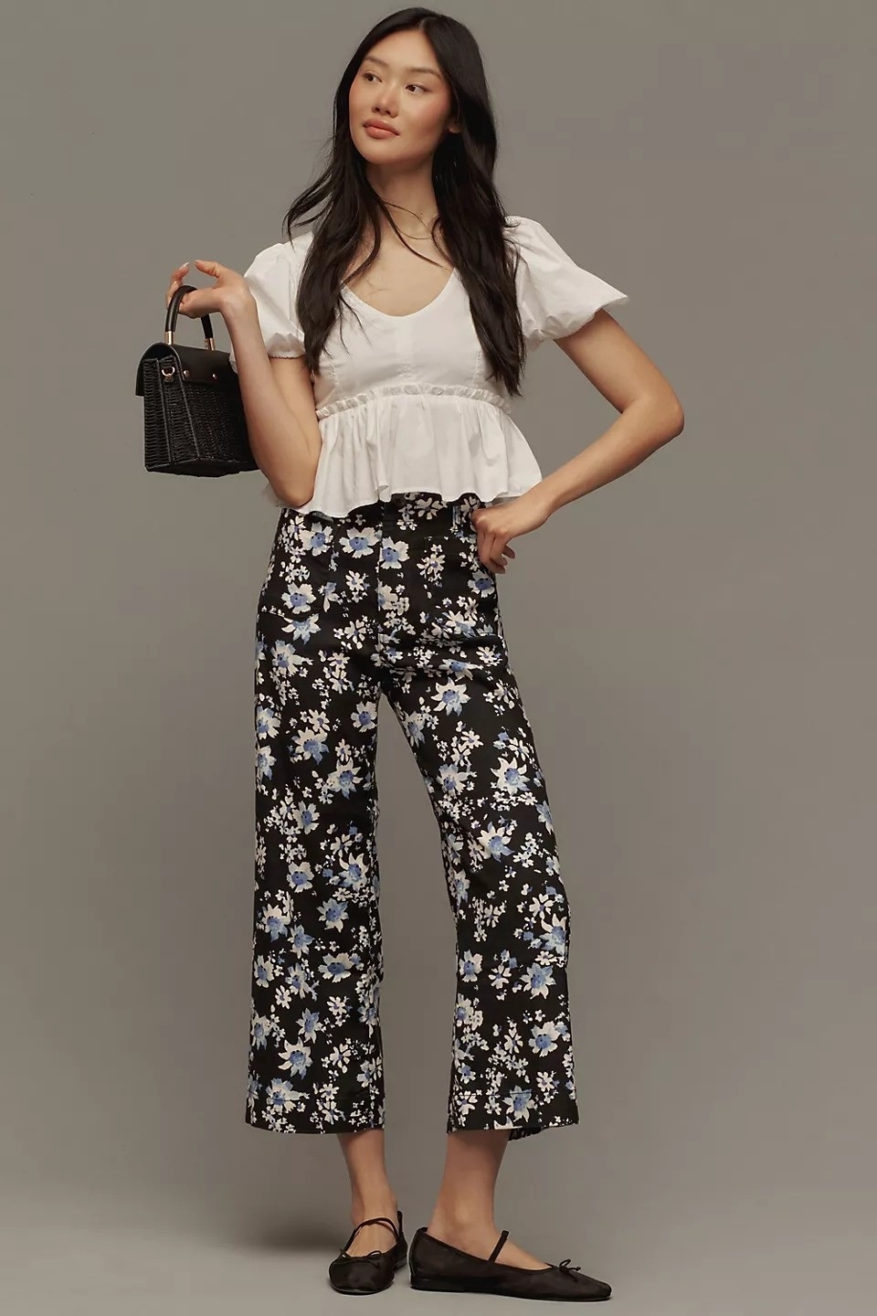 Woman posing in a ruffled white blouse, floral pants, and black flats, holding a small purse
