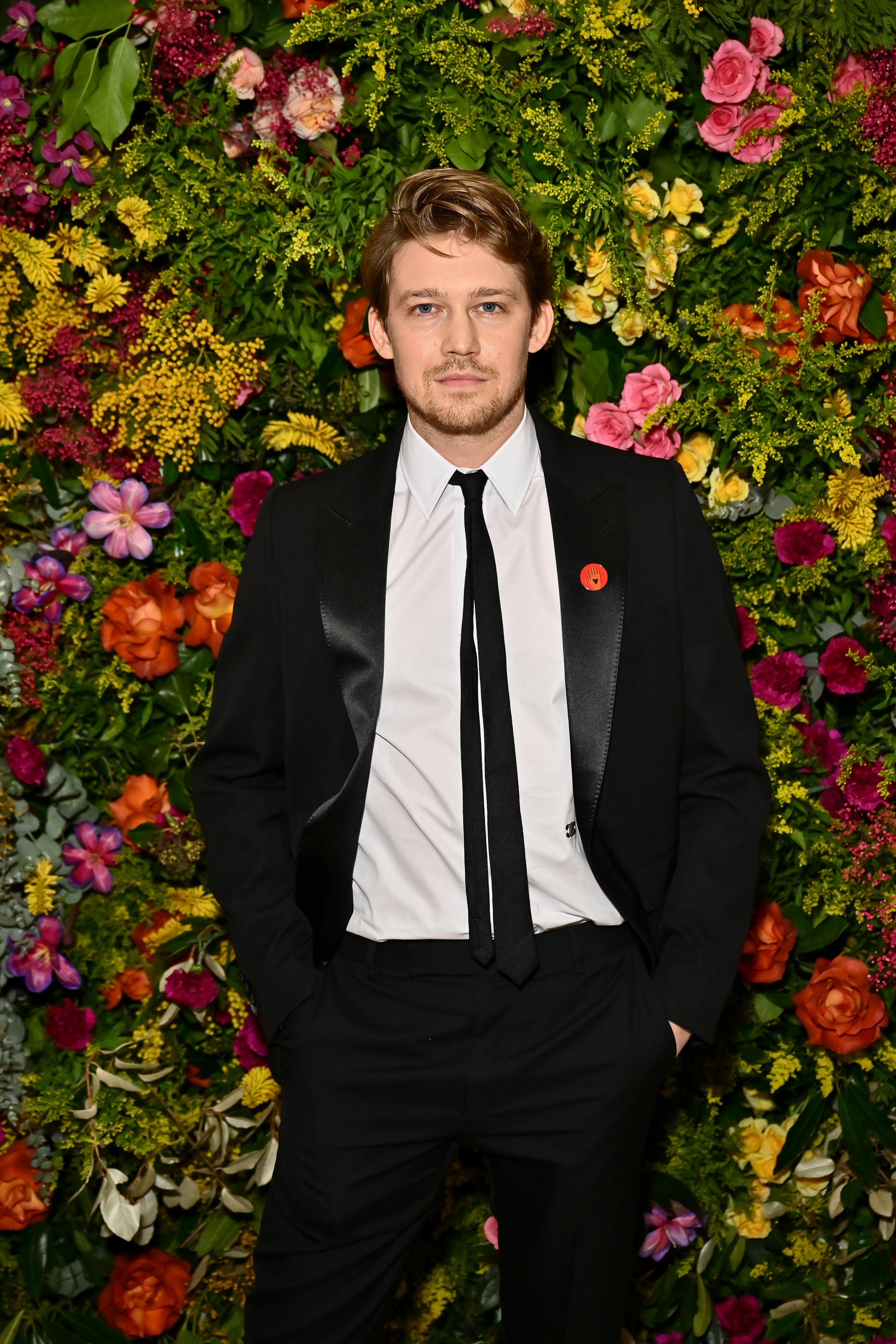Joe Alwyn standing in front of a floral backdrop, wearing a black suit and tie with a red lapel pin